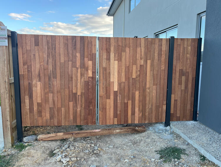 Merbau double gates with Monument steel frame installed beside a new home in Officer. Custom Merbau double driveway gates with powder-coated Monument steel frame completed in Officer to complement existing timber fencing and provide secure side property access.